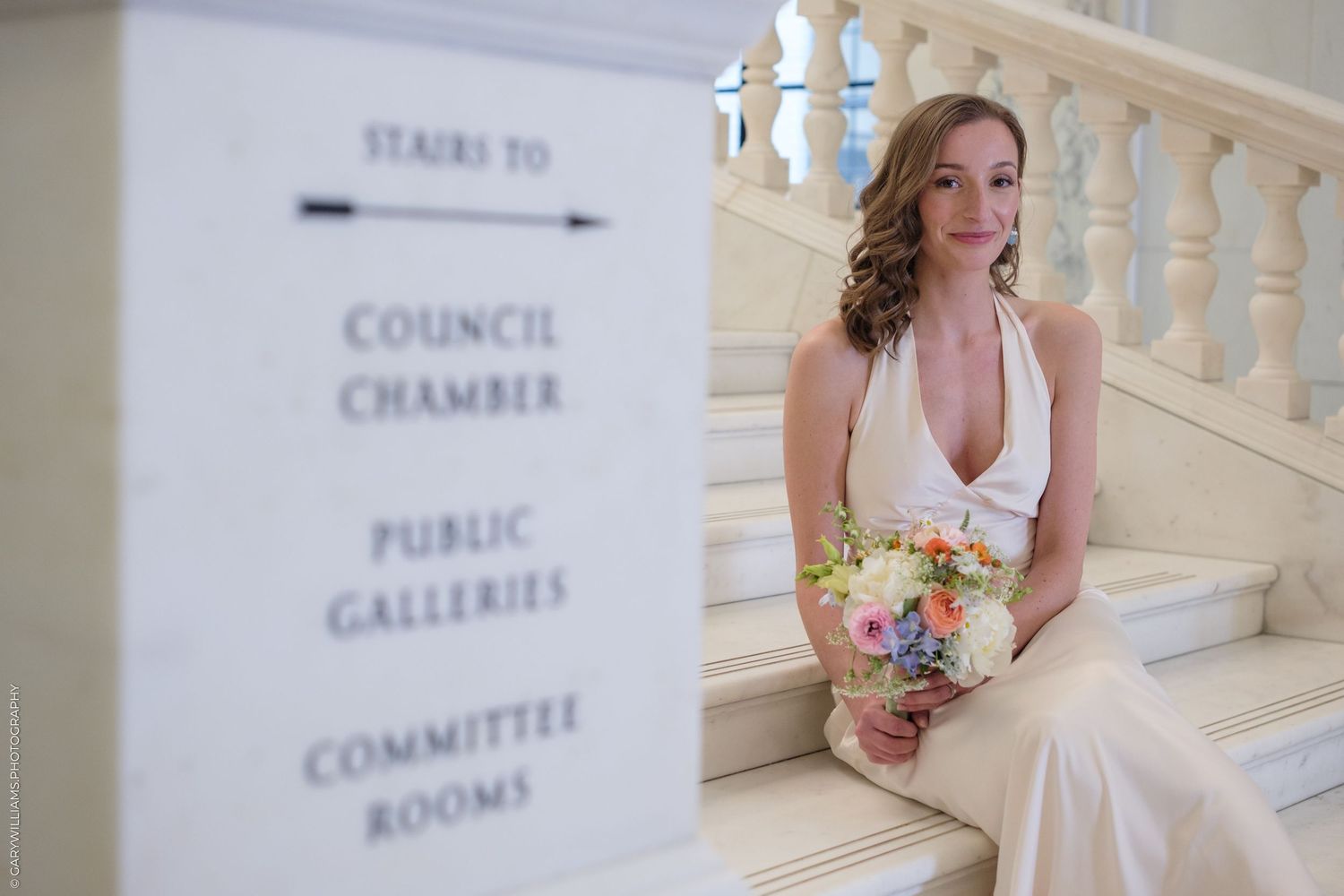 Bride in white dress stands by marble staircase near council chamber directory sign holding floral bouquet.