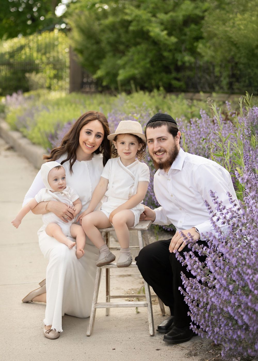 A family of four poses together on a bench next to blooming purple lavender flowers in a garden setting.