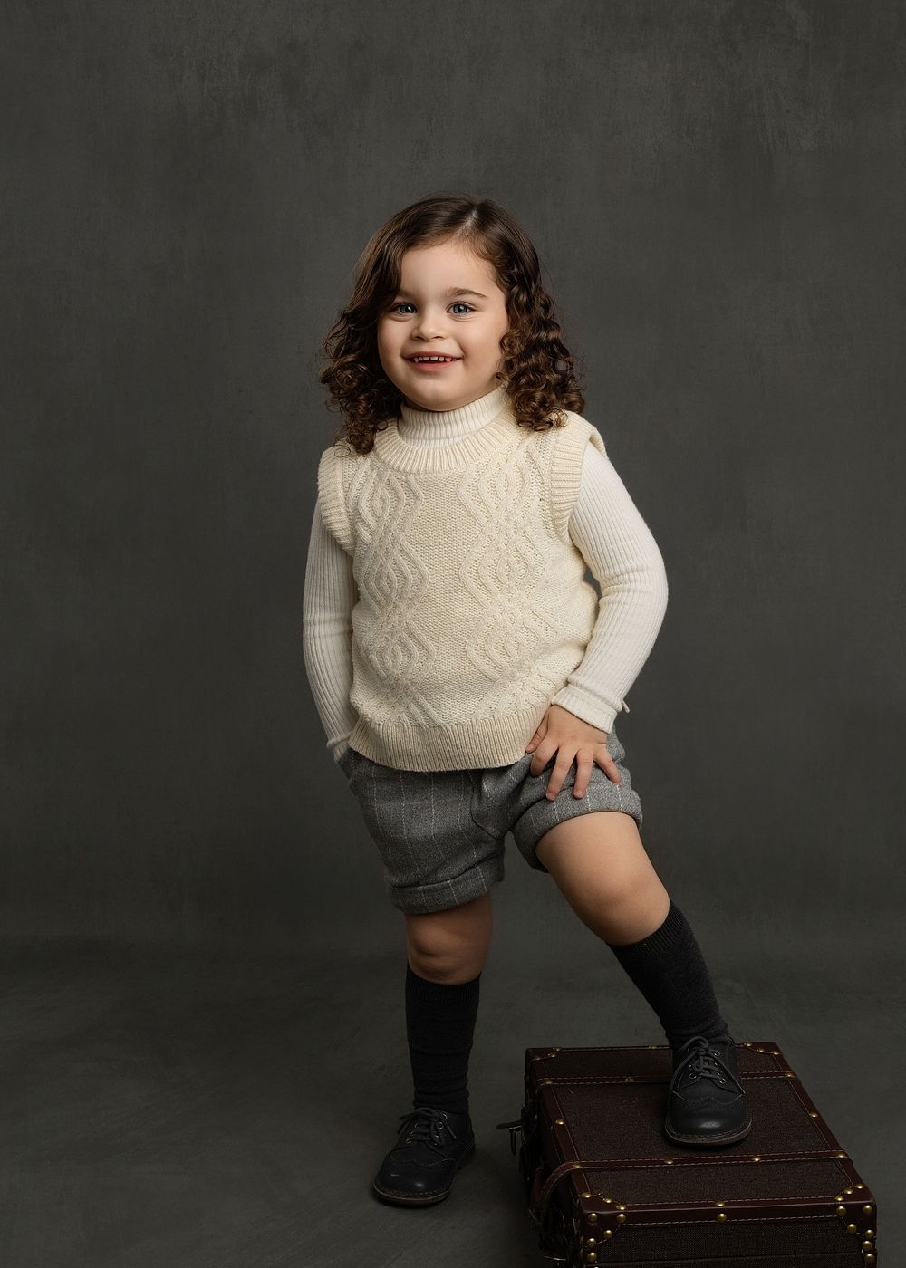 Child wearing cream cable knit sweater and shorts poses on vintage suitcase against dark backdrop in studio portrait.