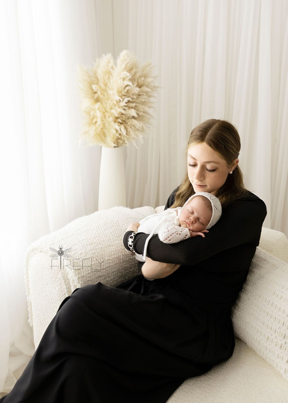 Mother in black dress cuddles newborn on white couch with pampas grass decor.