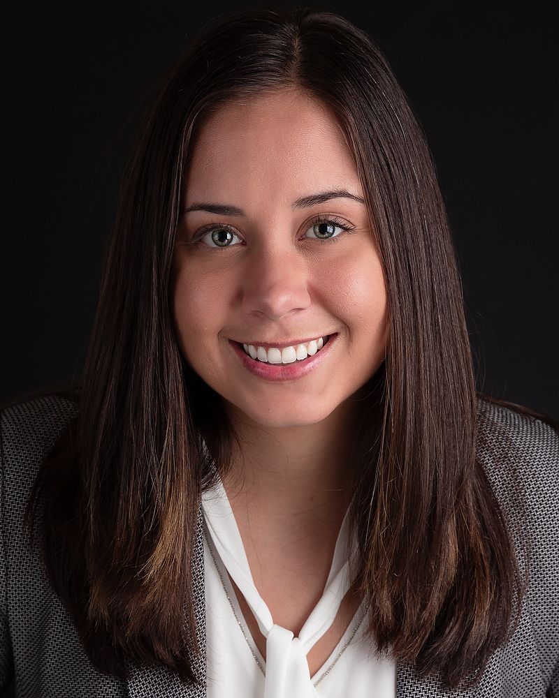 Professional headshot portrait with dark background showing a warm smile and long brown hair wearing a blazer and white blouse.