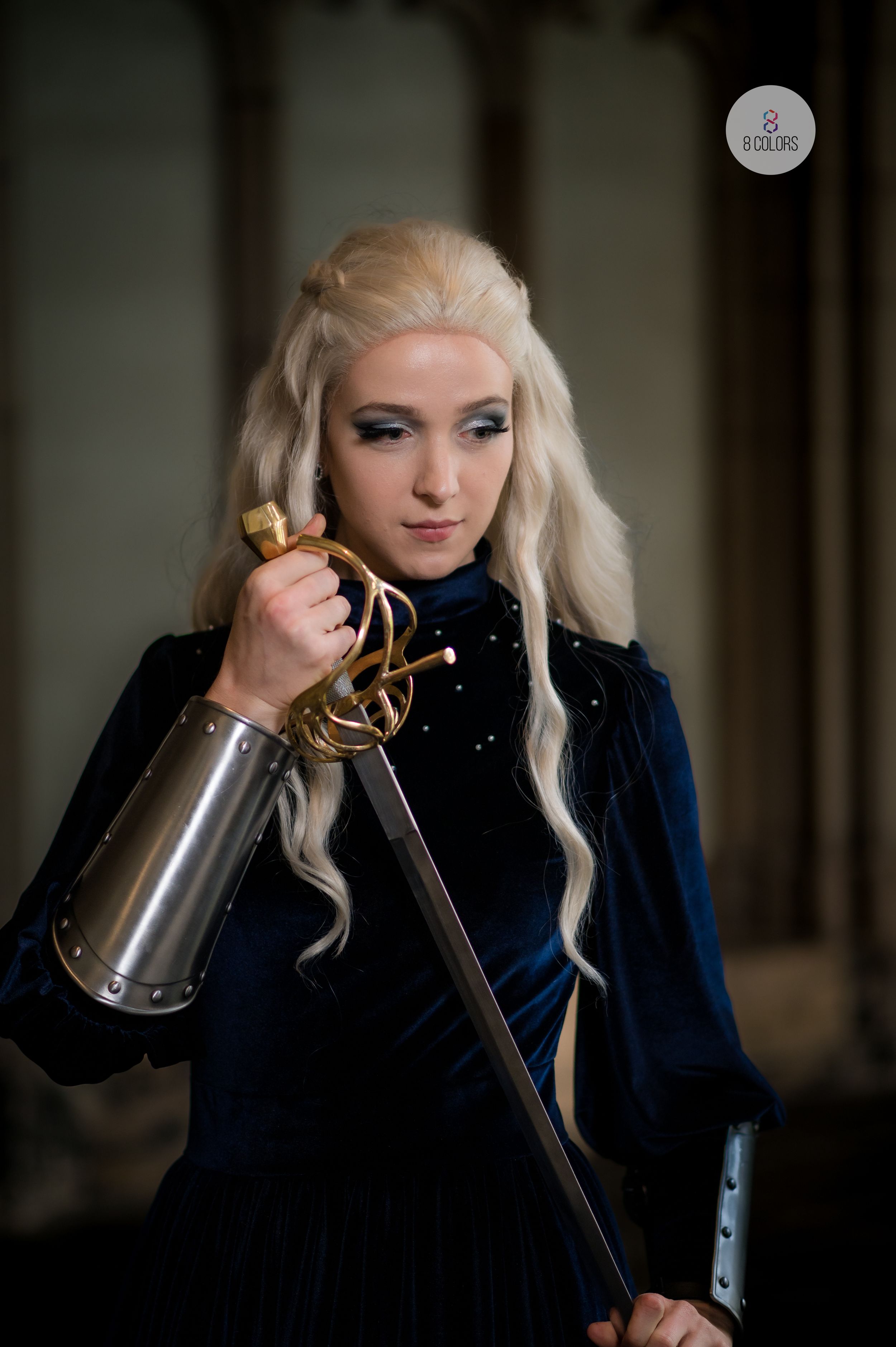 A cosplayer in a dark blue velvet gown holds a decorative sword while wearing long platinum blonde braided hair.