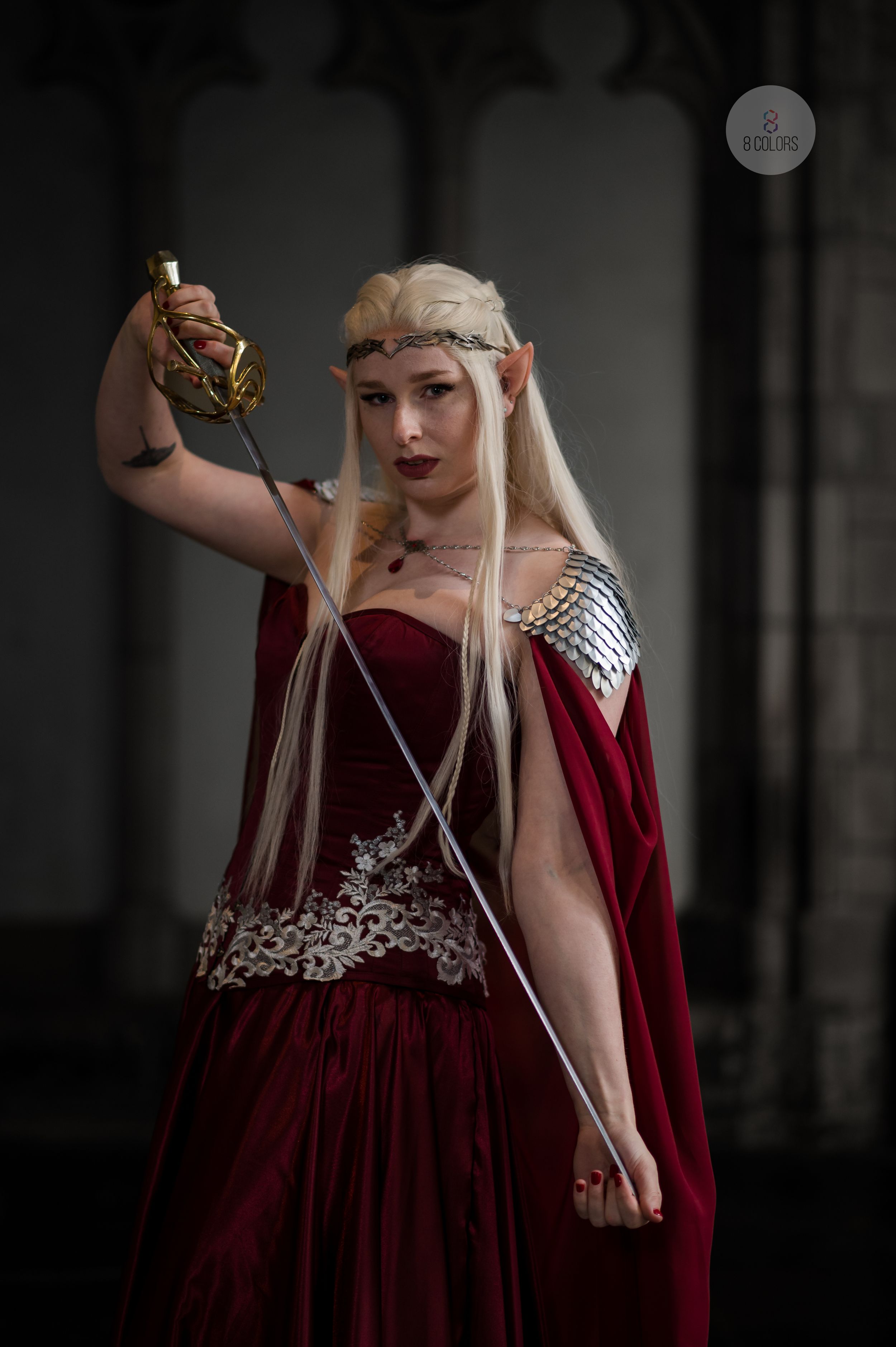 Fantasy cosplay model in red velvet medieval gown with silver chainmail armor and crown holding sword aloft.