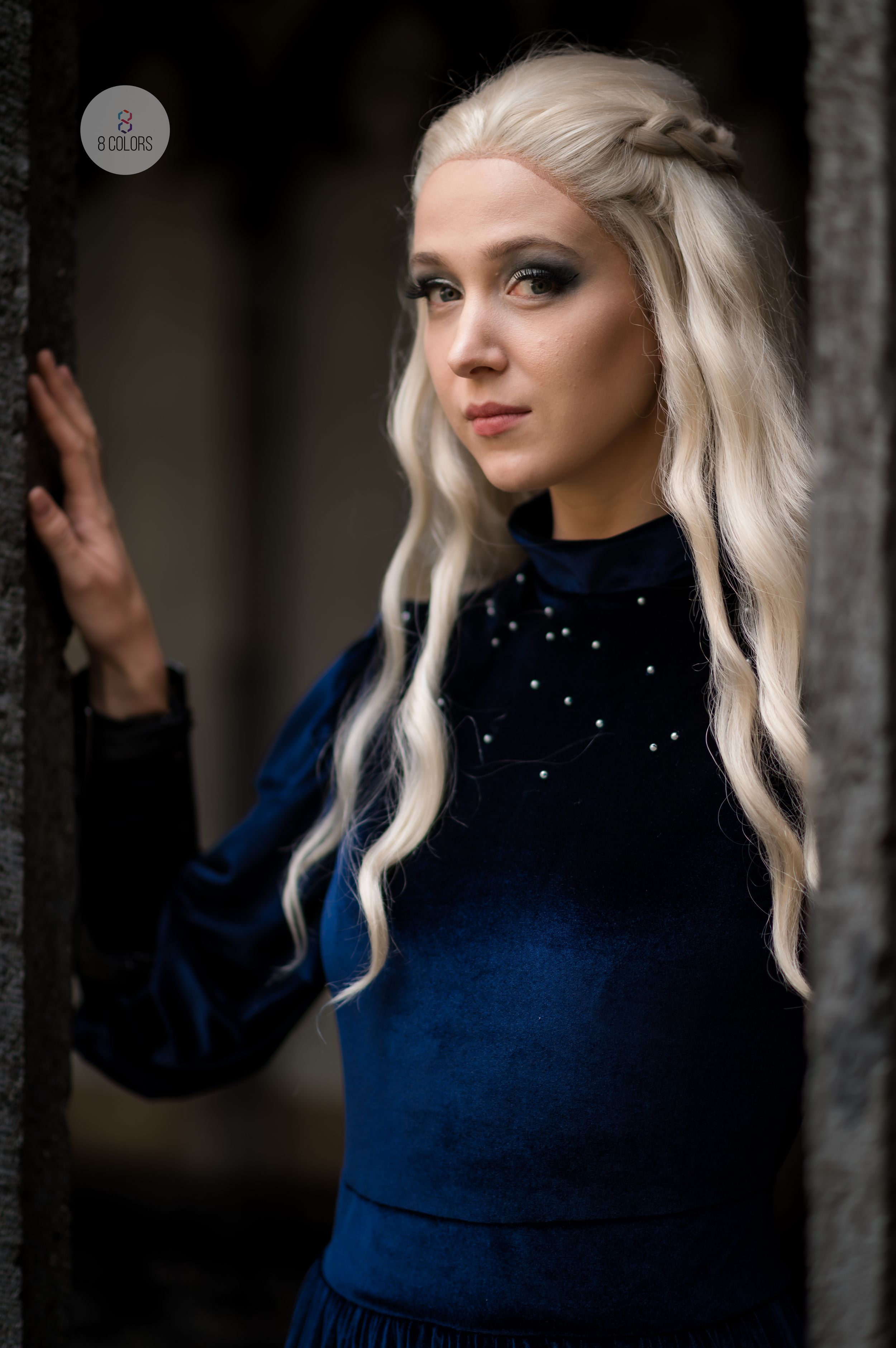 Portrait of woman with flowing platinum blonde hair wearing a dark blue sweater with embellishments against stone wall backdrop.