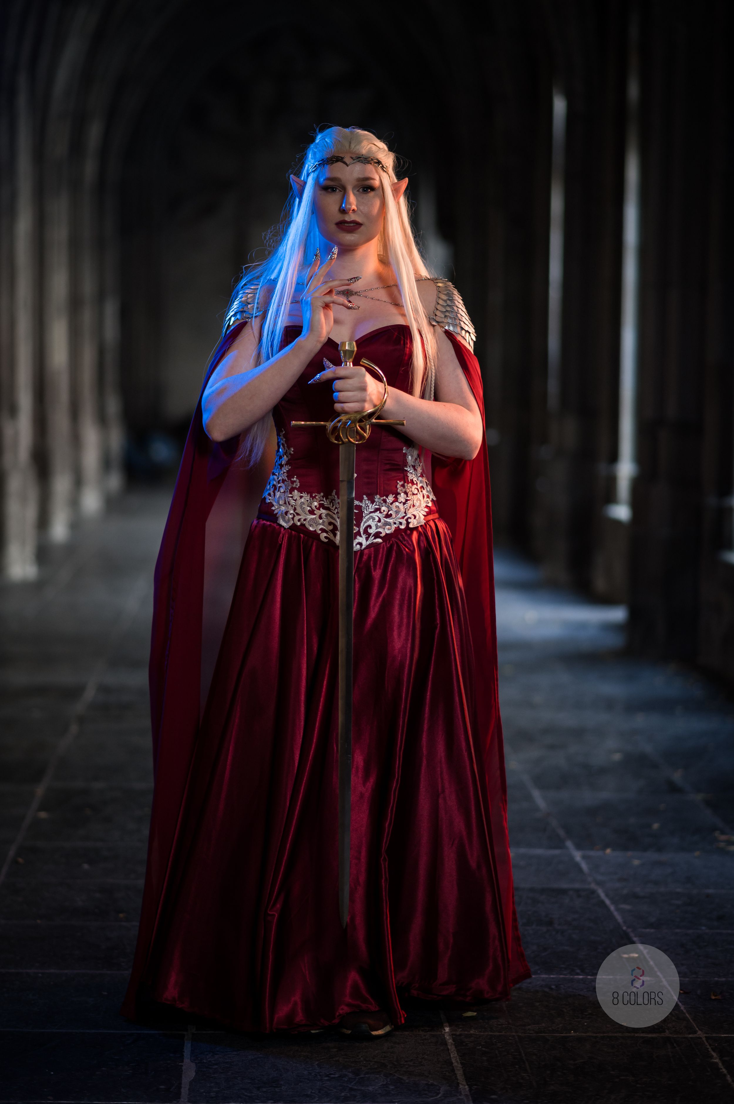 A cosplayer in a medieval-style burgundy dress and cloak stands in a dimly lit stone corridor holding a prop sword.