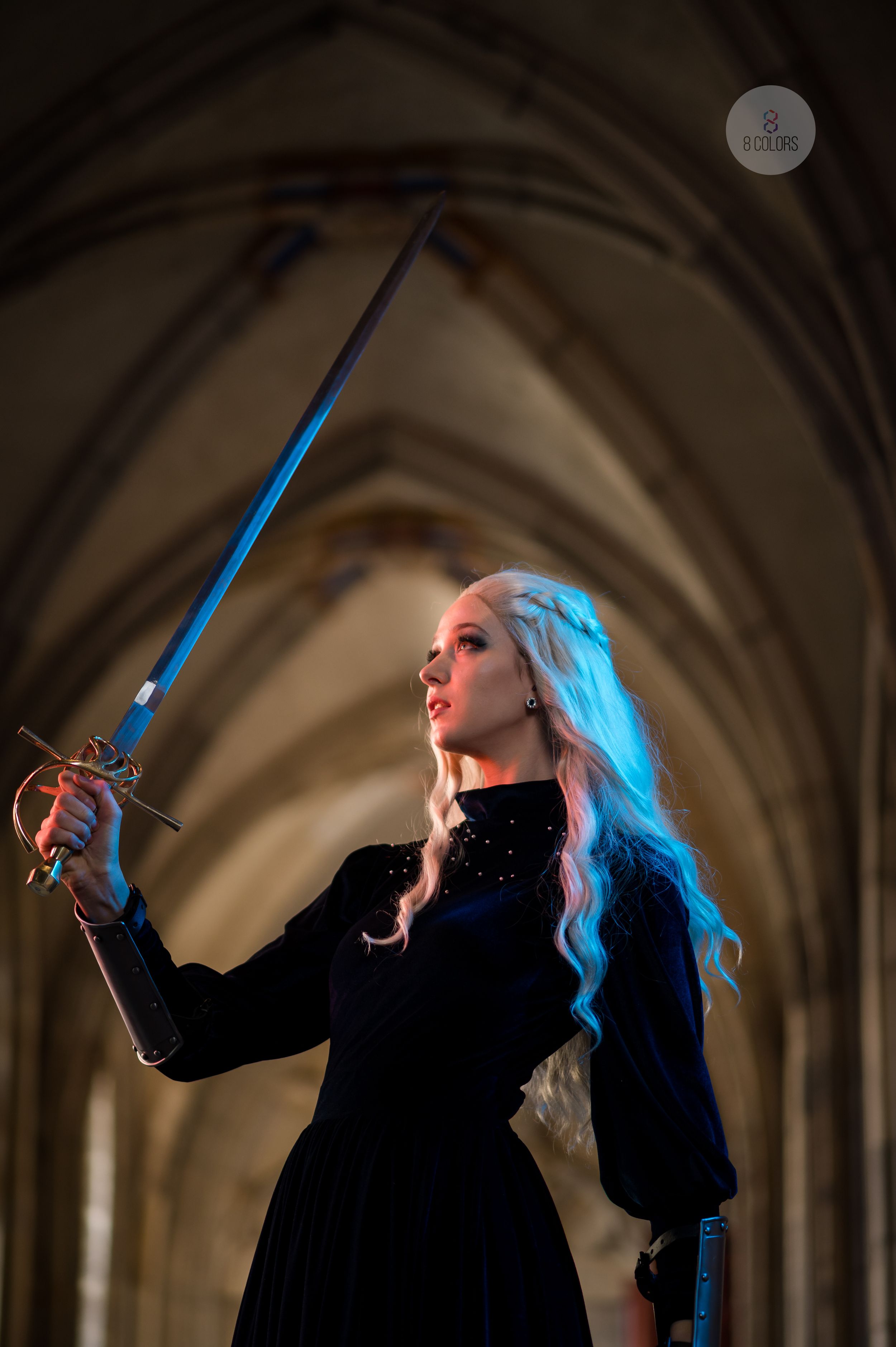 A figure in a black dress raises a glowing blue sword against a gothic vaulted cathedral ceiling.