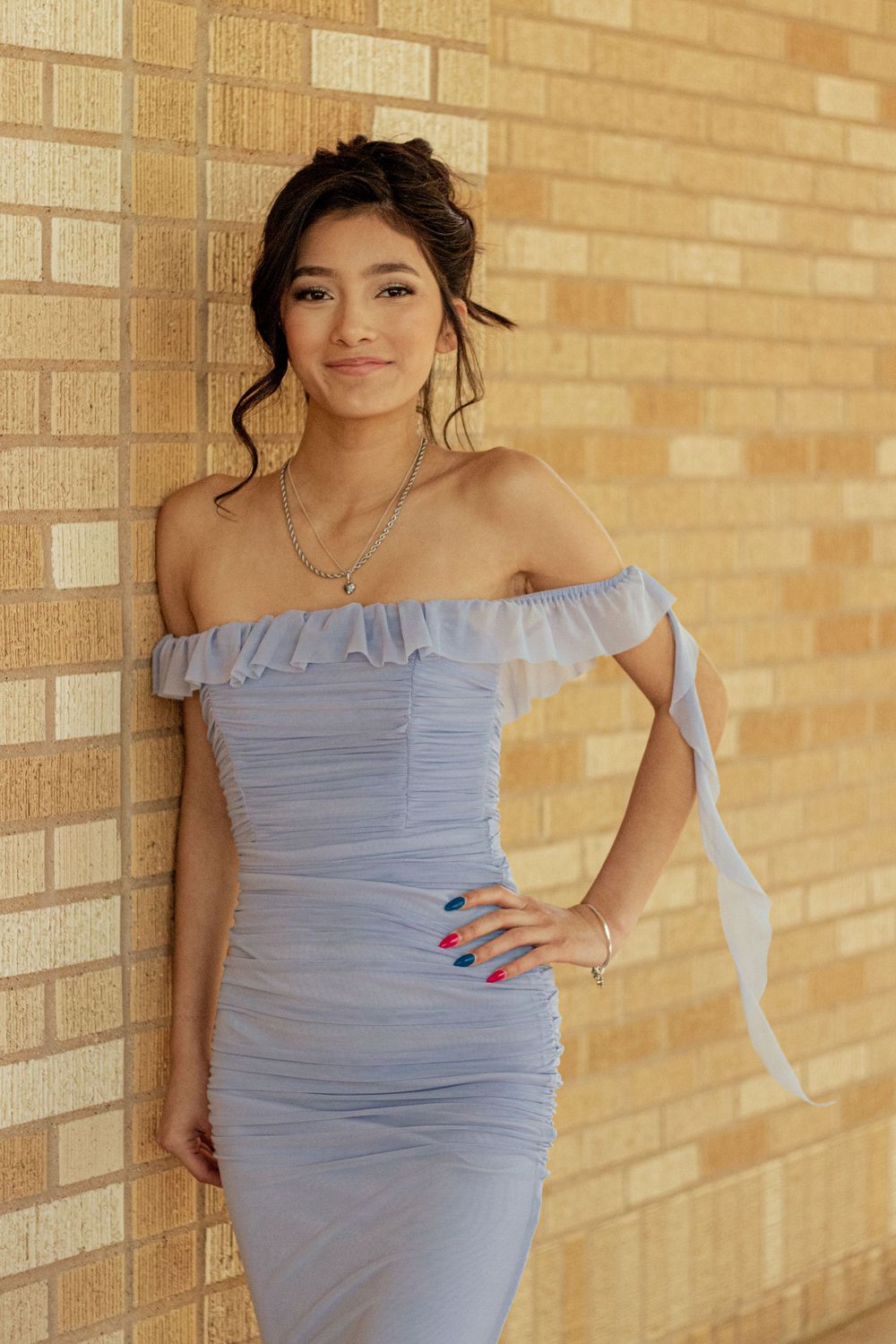 A person in a light blue off-shoulder ruched dress poses against a brick wall in warm lighting.