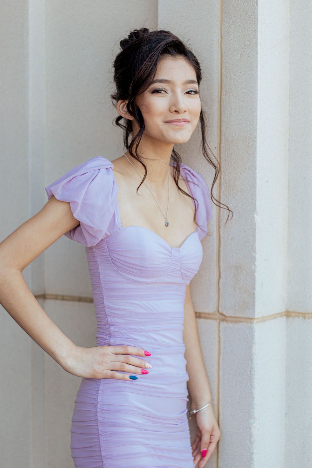 Elegant woman in a form-fitting lavender dress with ruffled cap sleeves poses against a light colored wall.
