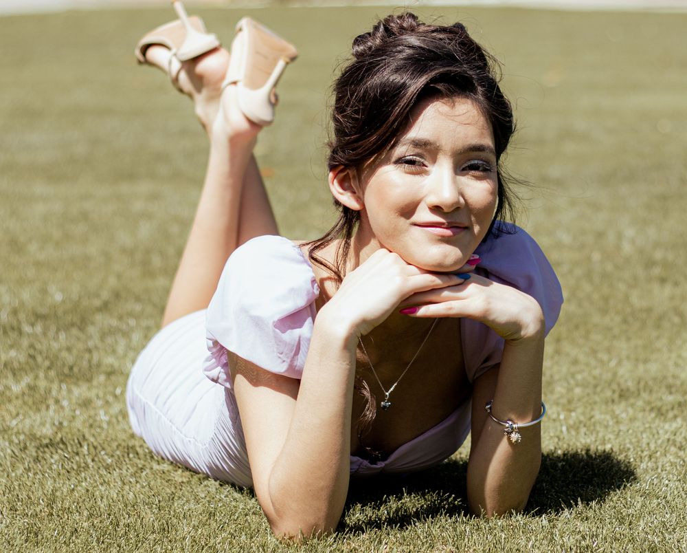 A person in a white dress lies on green grass in a sunny meadow, resting their chin on folded hands.