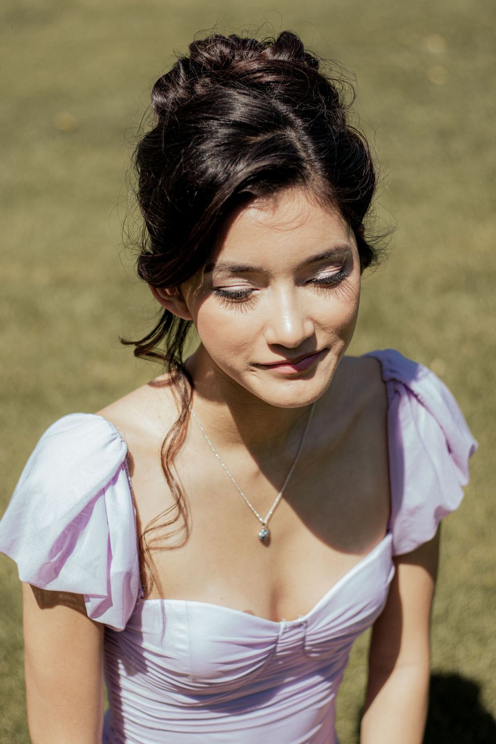 A person in a romantic lavender dress with puff sleeves and upswept dark hair gazes down while wearing a delicate necklace.