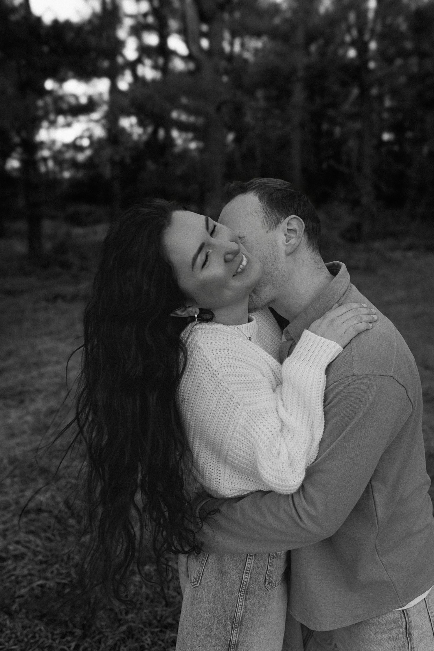 A romantic black and white photo capturing an intimate embrace between a couple laughing together outdoors.