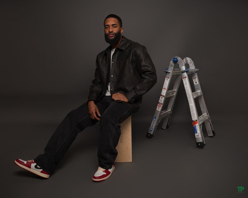 Portrait of person in dark denim outfit and red sneakers seated next to silver metal ladder against dark background.