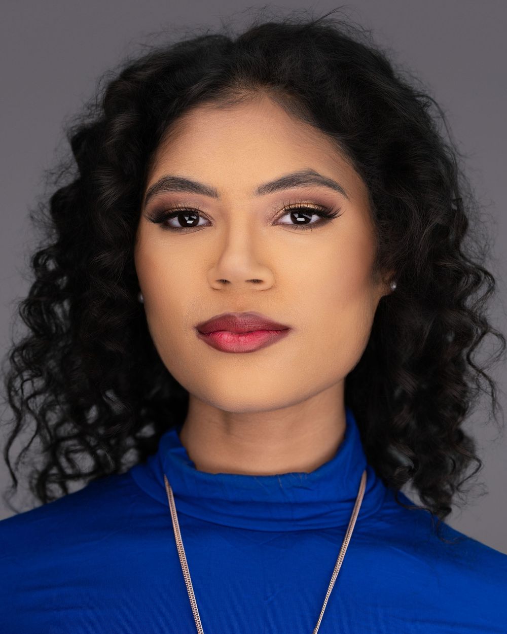 Portrait of a woman wearing a bright blue turtleneck and necklace, with natural curly hair and glamorous makeup.