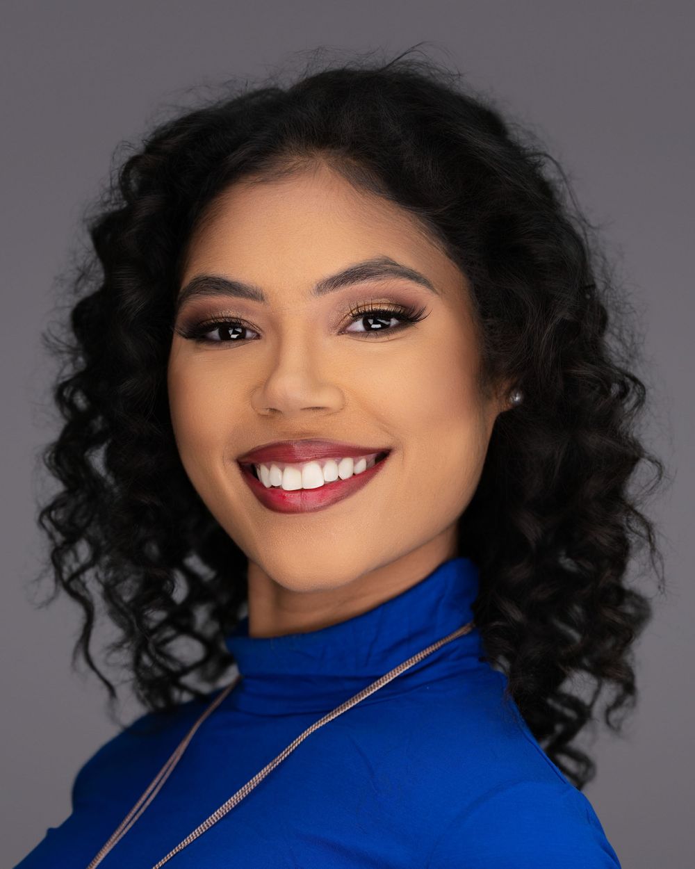 Professional headshot of a person in a royal blue turtleneck against a gray backdrop, with curly hair and bright smile.