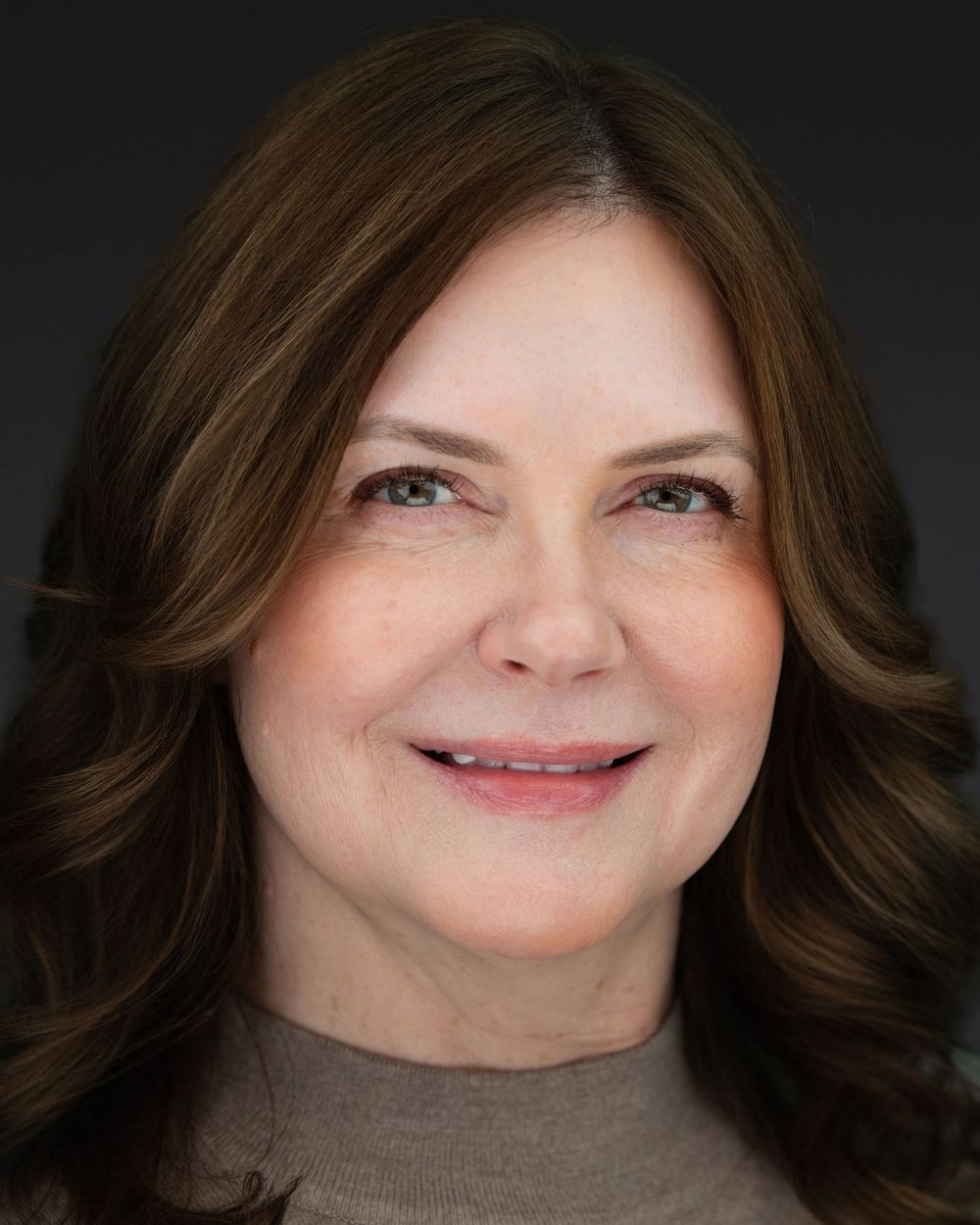 Professional headshot portrait of a woman with long brown wavy hair wearing a taupe sweater against dark background.