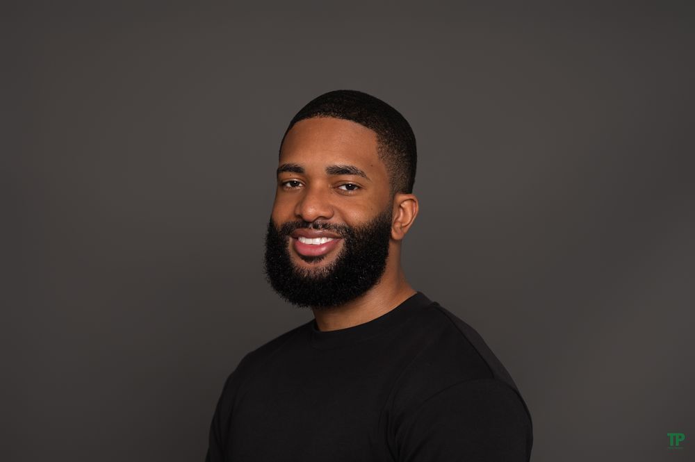 Professional portrait of a smiling person with beard wearing black shirt against dark gray background.