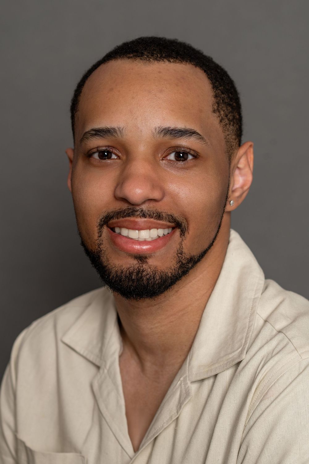 Professional headshot portrait of someone smiling while wearing a beige collared shirt against a dark gray background.