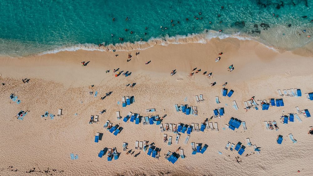 Aerial view of a sandy beach with blue umbrellas and lounge chairs lined up along the shore.