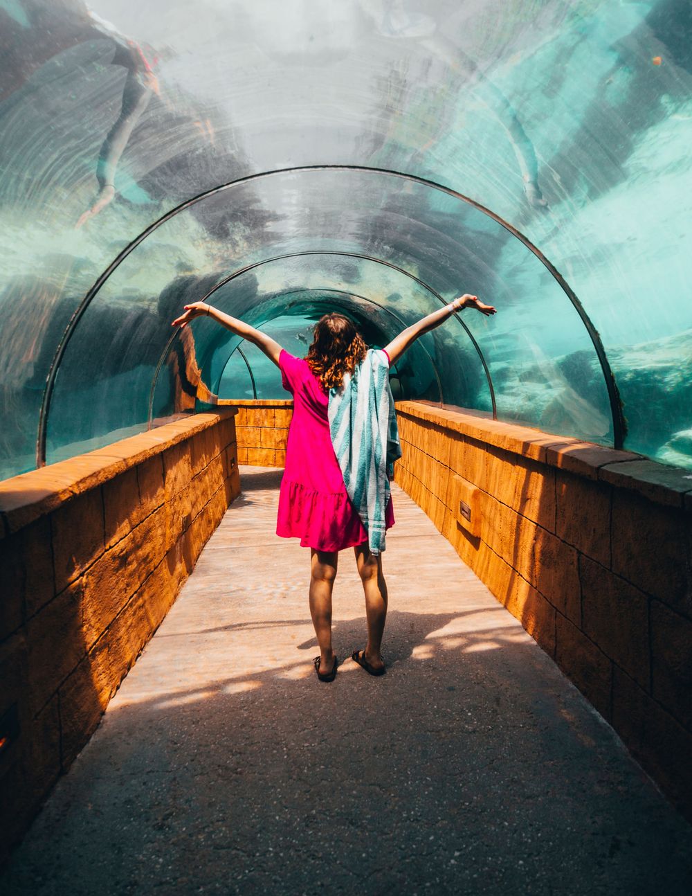 Figure in pink dress standing in underwater tunnel walkway with arms outstretched.