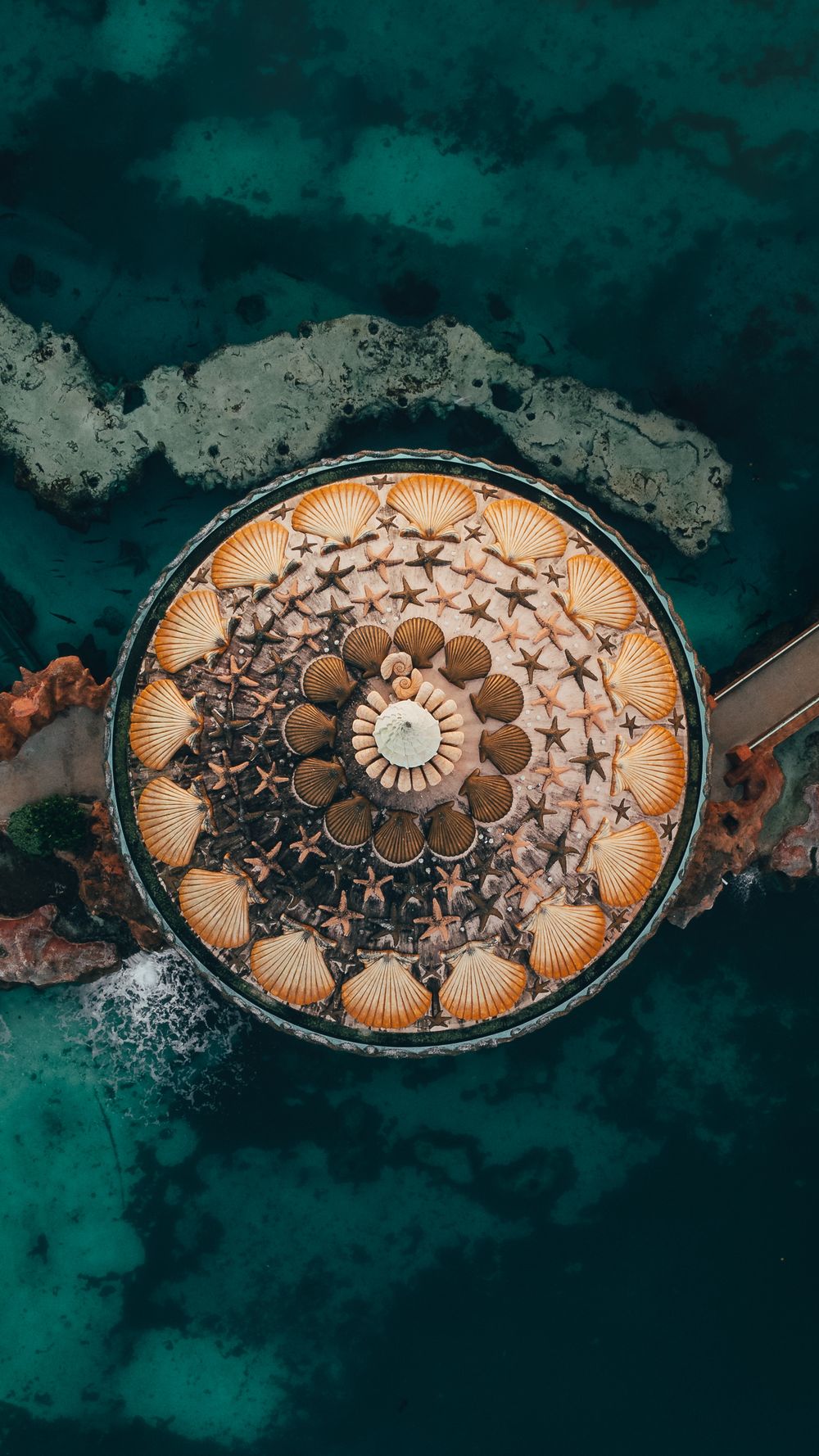 Ornate circular mosaic pattern with seashell designs photographed against turquoise ocean water.