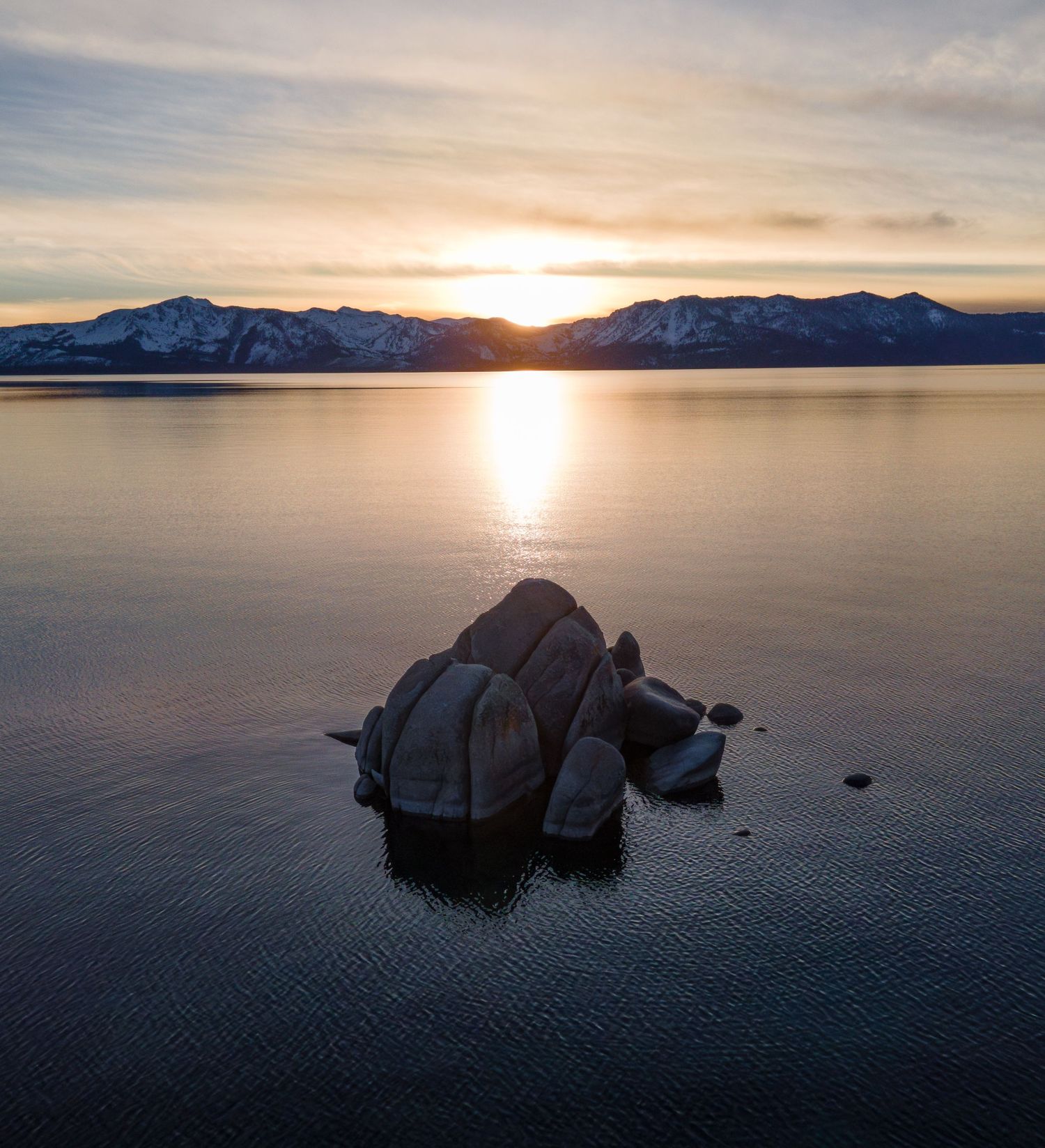 Aerial view of Bonsai Rock in Lake Tahoe at sunset with snow-capped mountains in the background and golden light reflection.