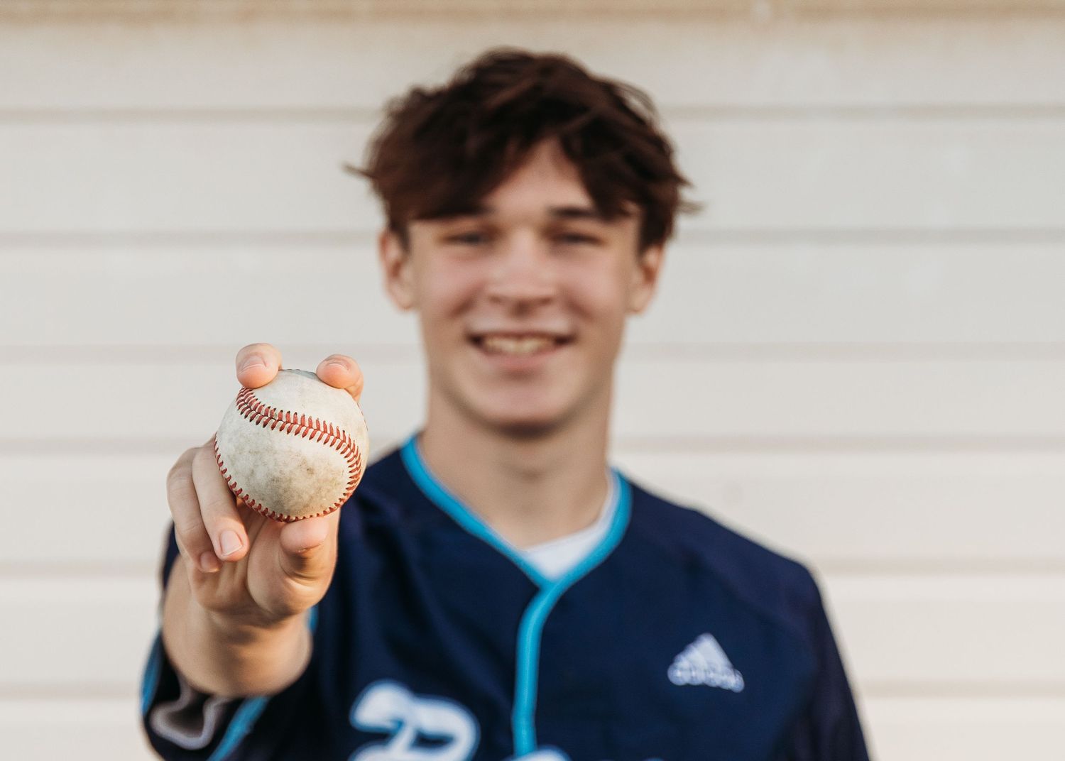 Baseball player wearing a navy blue jersey holds up a baseball while standing against a light colored wall.