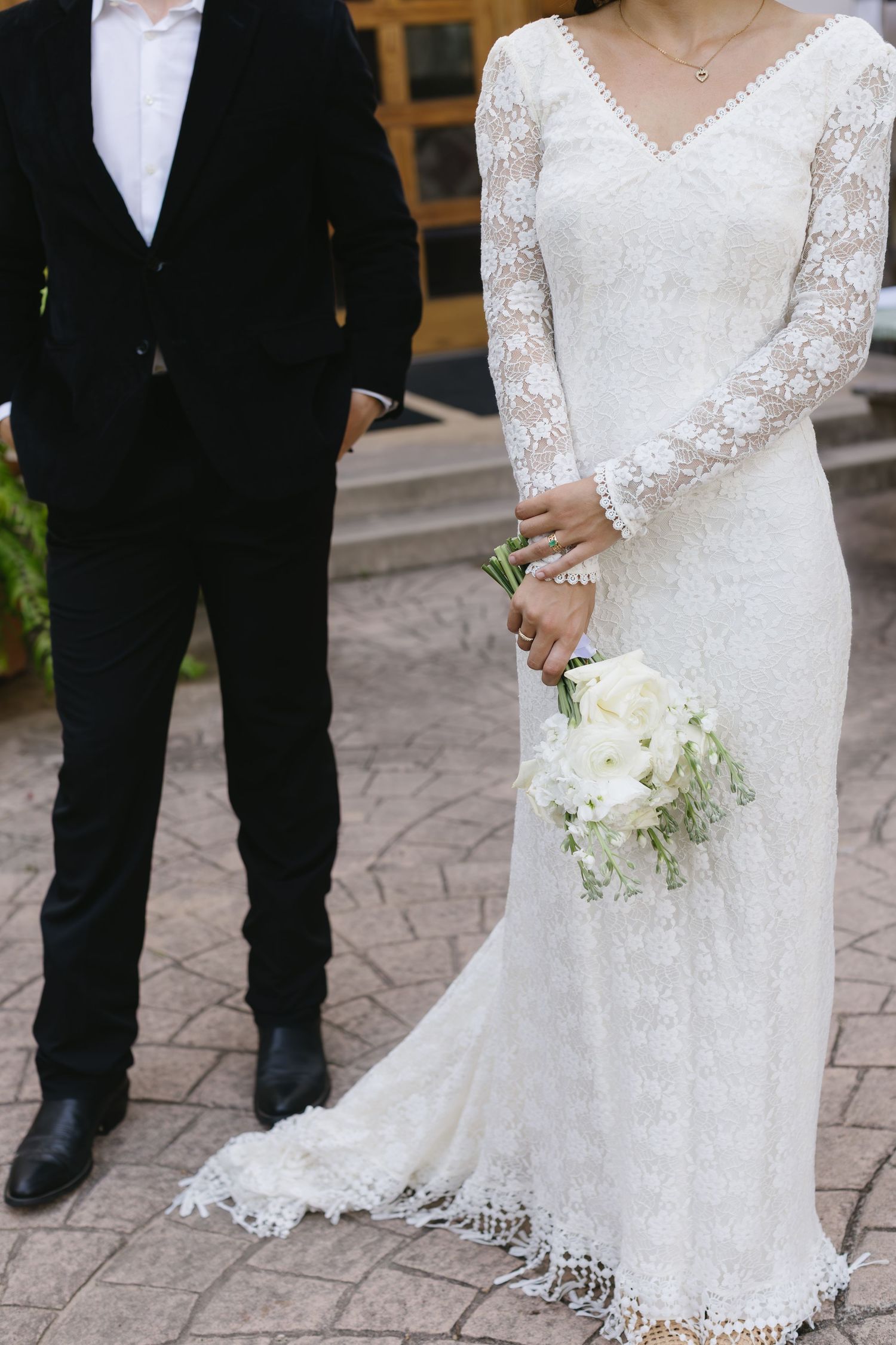 A bride in a sleek white dress and groom in tuxedo walk down a brick pathway during an outdoor garden ceremony.