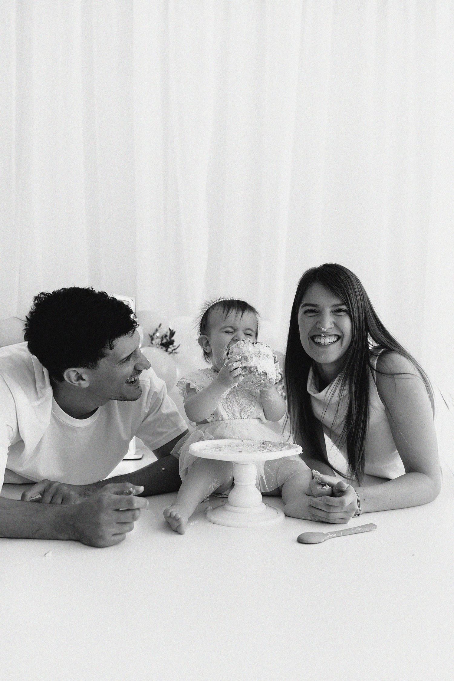 Black and white photo of a family lying together and smiling while their baby plays with a cake on a white cake stand.
