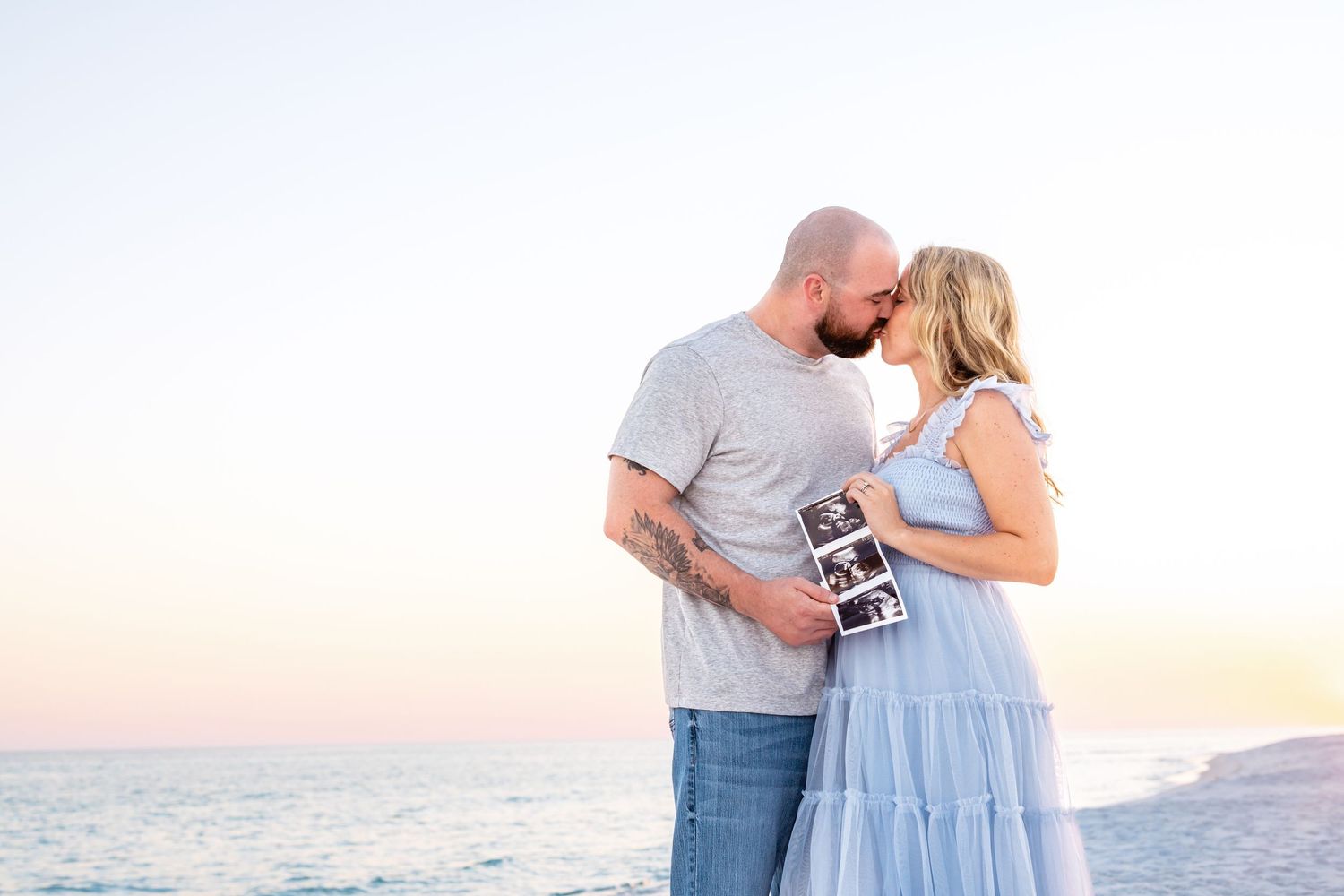 A couple standing together on a beach at sunset holds an ultrasound photo while sharing an intimate moment.
