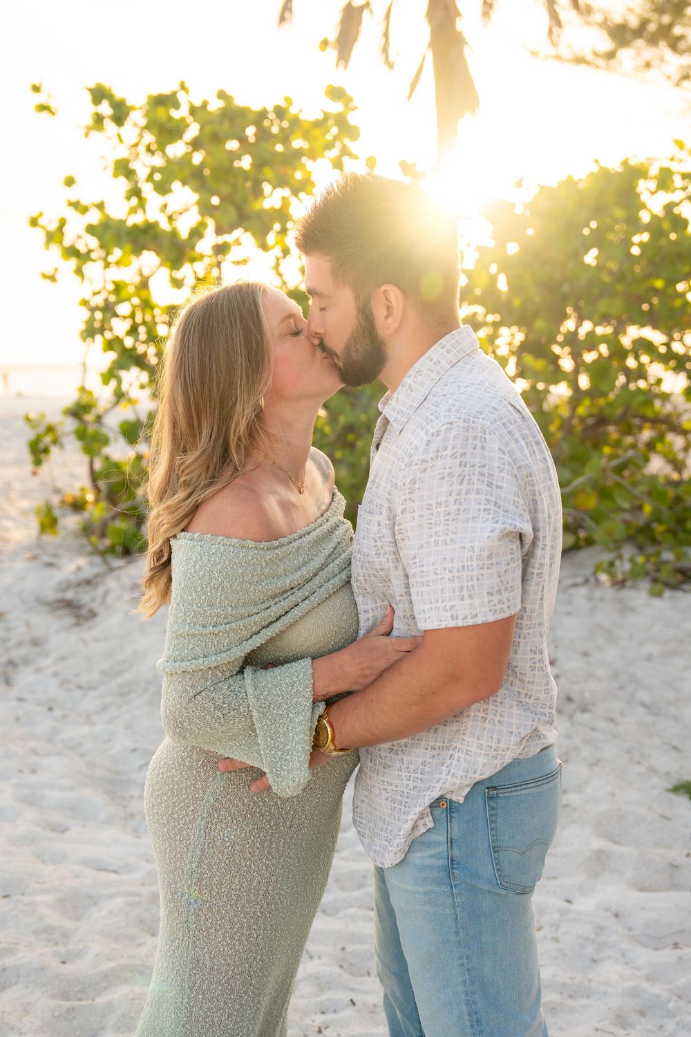 Couple sharing a romantic kiss on a tropical beach at sunset during a maternity photo session.