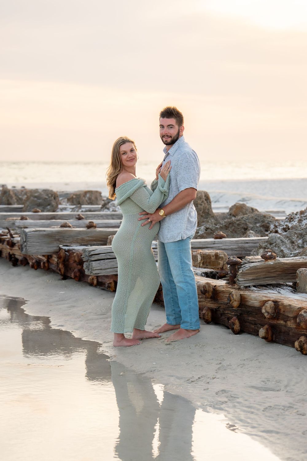 Smiling pregnant woman in mint dress leans into partner's embrace on sandy beach at warm sunset.