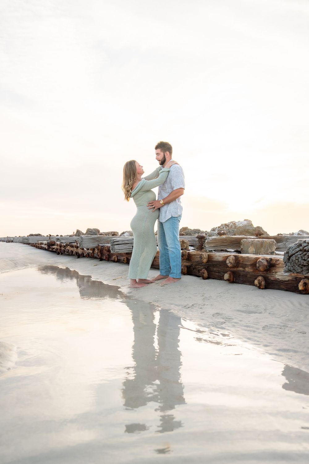 Pregnant couple facing each other tenderly on reflective wet sand beach at golden hour by rock jetty.