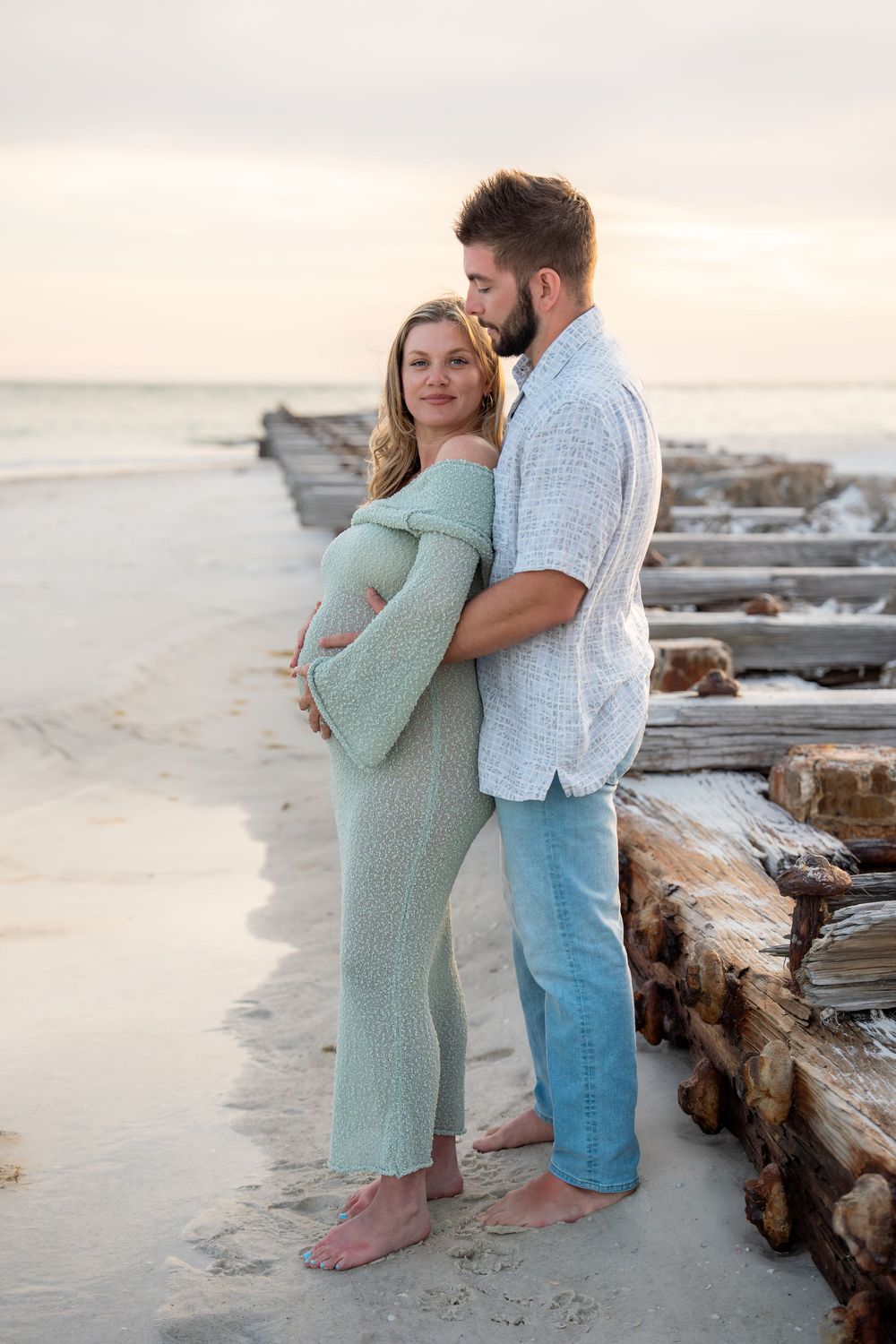 Couple sharing tender moment on beach at sunset, man kissing pregnant wife.