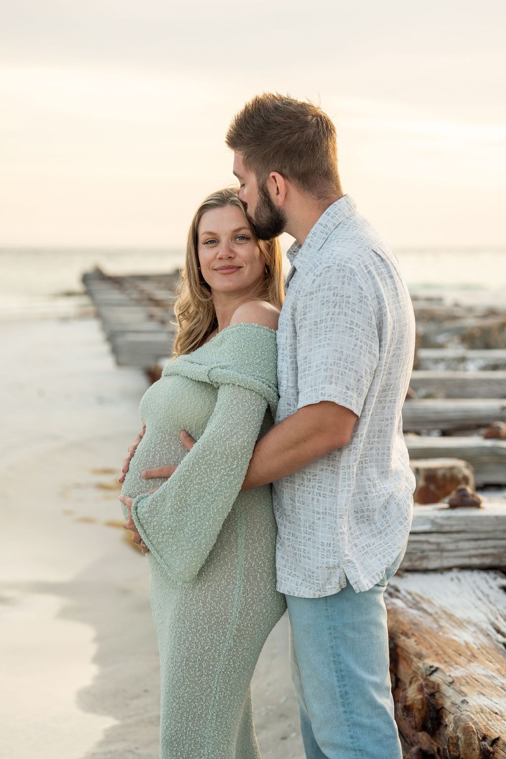Man kissing pregnant woman's temple as she smiles, standing on beach at sunset near driftwood.