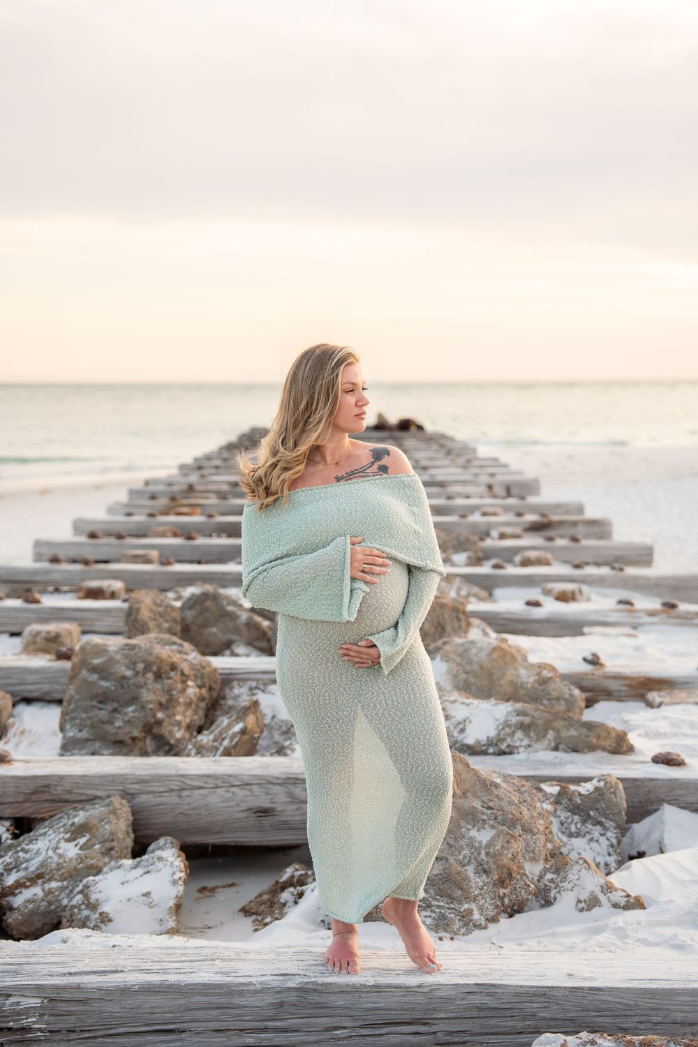 Pregnant woman in mint knit dress standing on beach pier ruins at sunset.