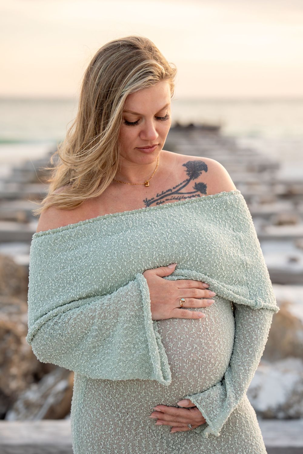 Blonde pregnant woman in mint off-shoulder knit dress cradling baby bump at beach.