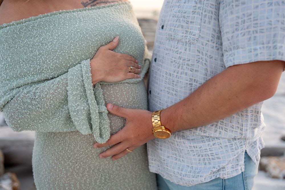 Close-up of couple touching pregnant belly, man wearing gold watch at beach.