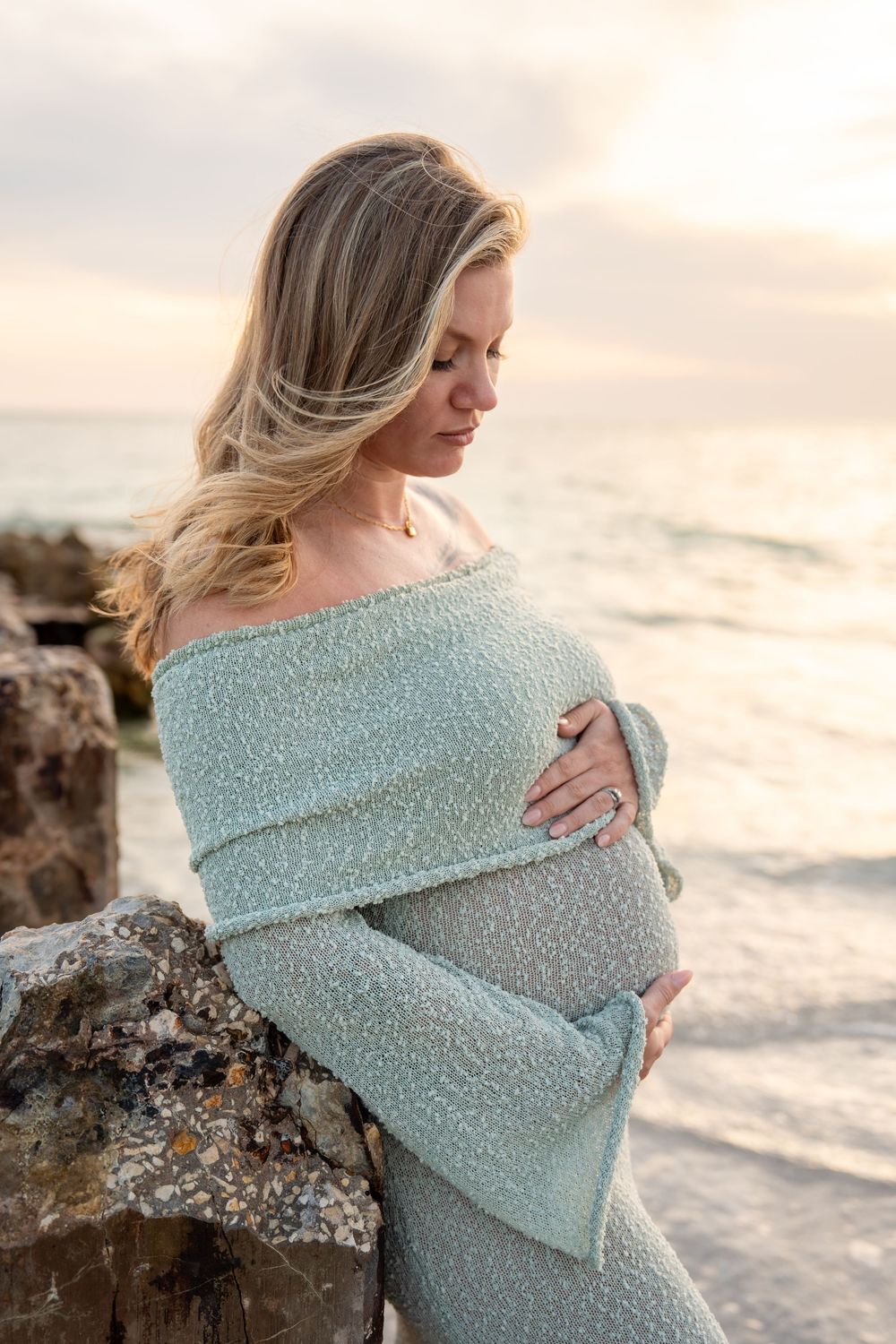 Pregnant blonde woman in mint knit dress holding belly near ocean rocks at sunset.
