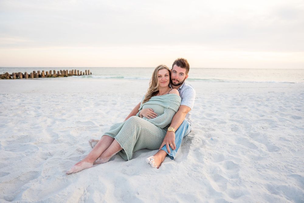 Couple sitting together on white sand beach at sunset, woman visibly pregnant.