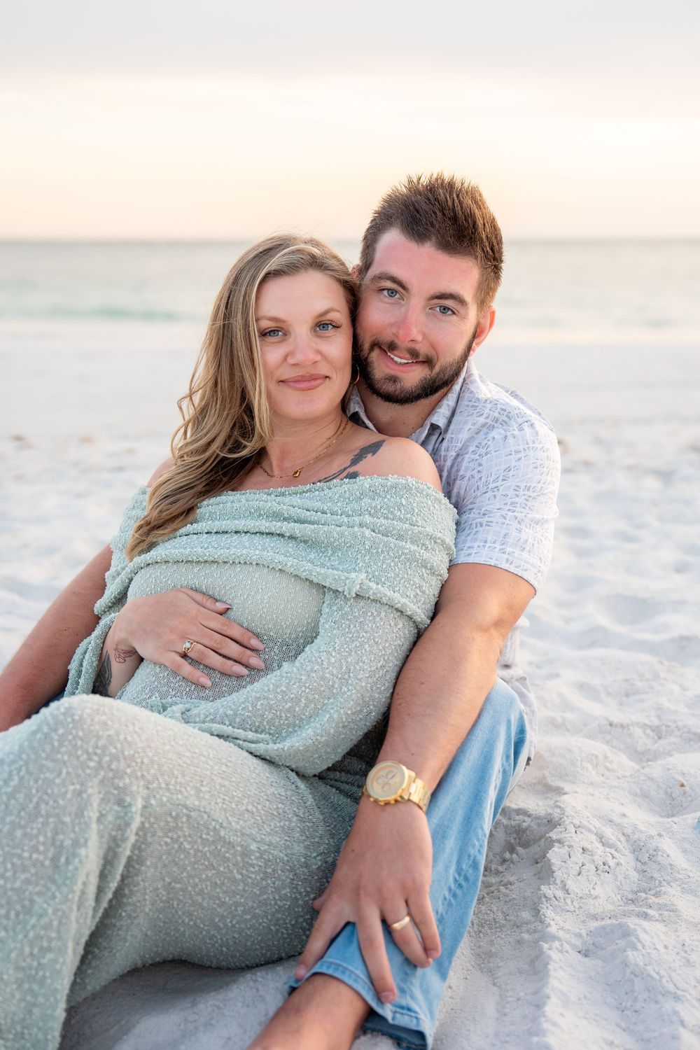 Smiling couple sitting on beach sand, pregnant woman cradled by partner at sunset.