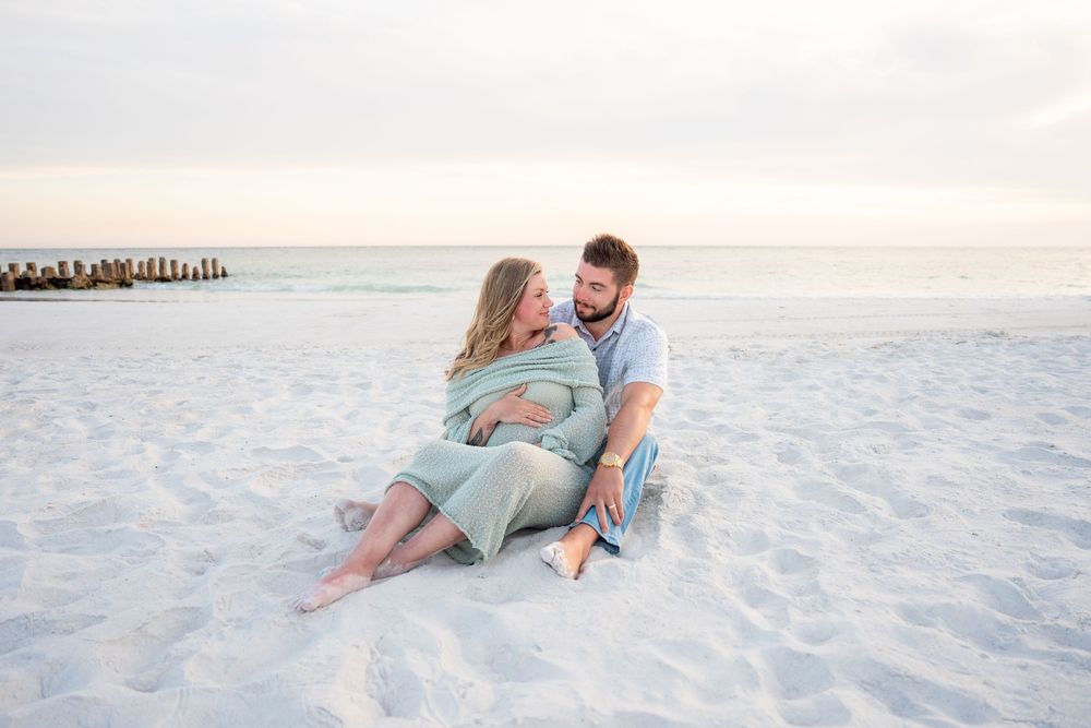 Couple sitting intimately on white sand beach at golden hour, woman pregnant.
