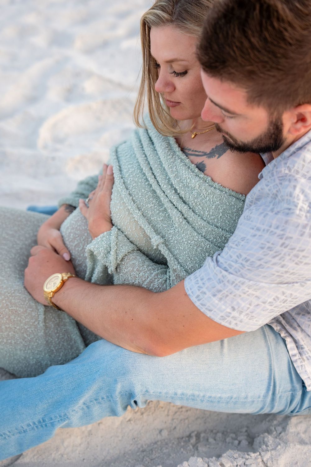 Couple sharing tender moment on sandy beach, man embracing pregnant woman wrapped in sage green knit blanket.