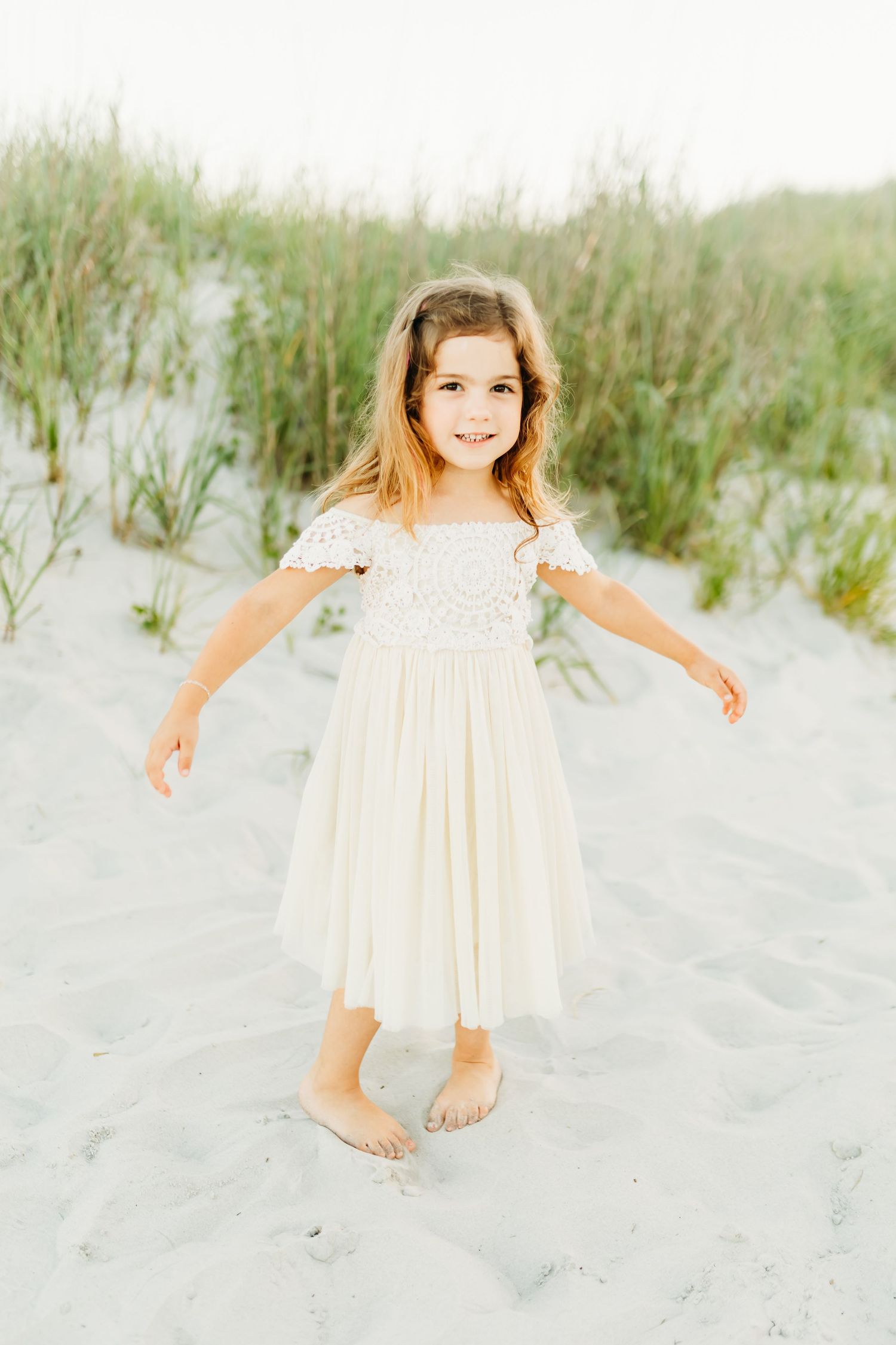 Young child wearing white lace dress stands barefoot on sandy beach with flowing grasses in background.