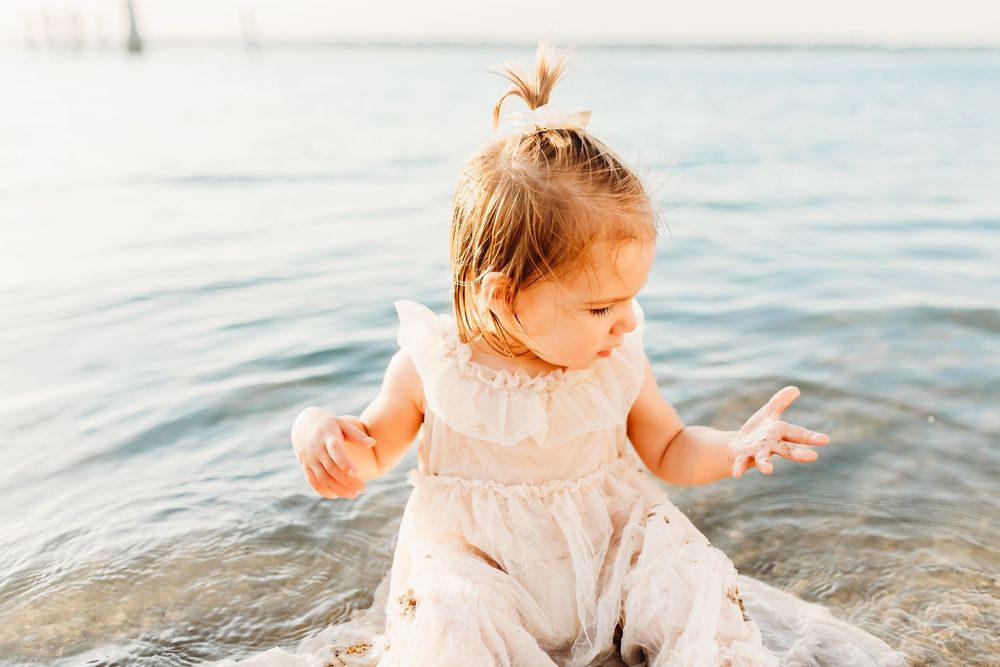 Small toddler in white dress sits and plays in shallow ocean water during golden hour.