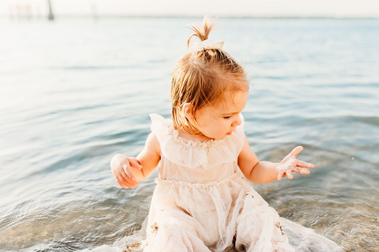 Small toddler in white dress sits and plays in shallow ocean water during golden hour.