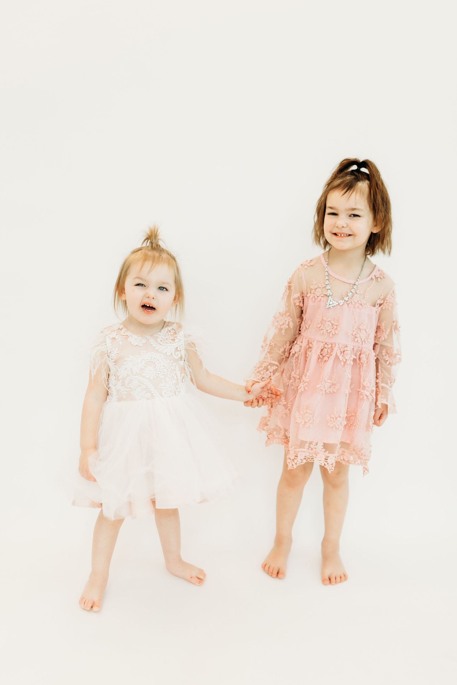 Two children in delicate lace dresses, one in white and one in pink, stand together holding hands on a light background.