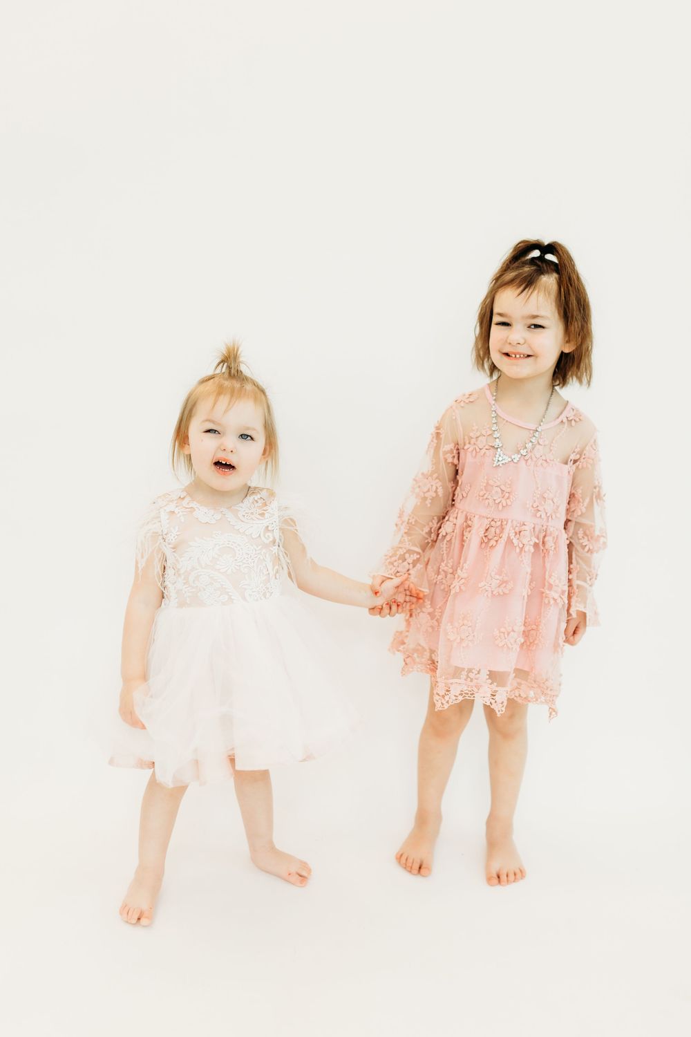 Two children in delicate lace dresses, one in white and one in pink, stand together holding hands on a light background.