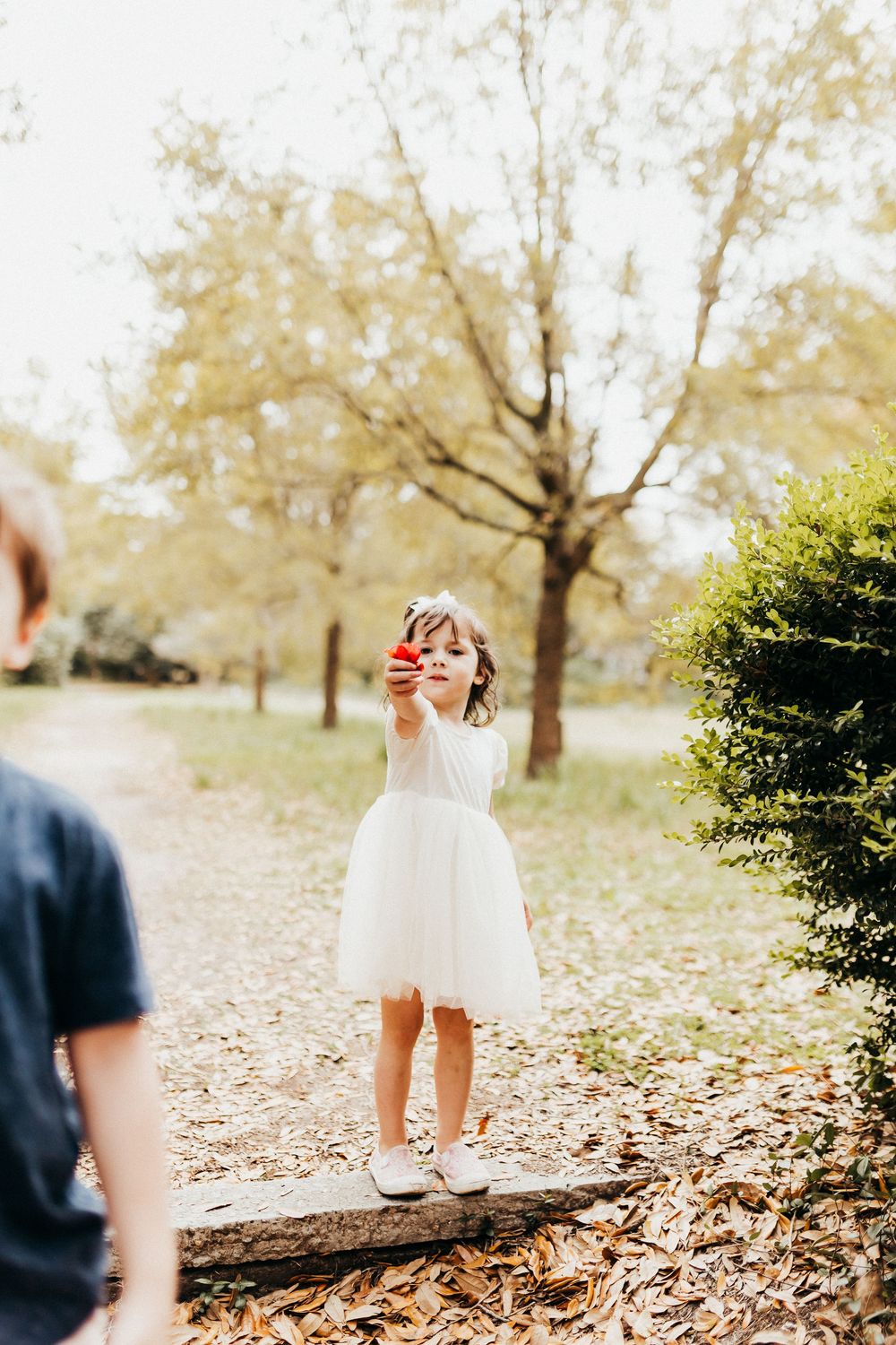 A young child in a white dress stands on fallen leaves in a park on an autumn day.
