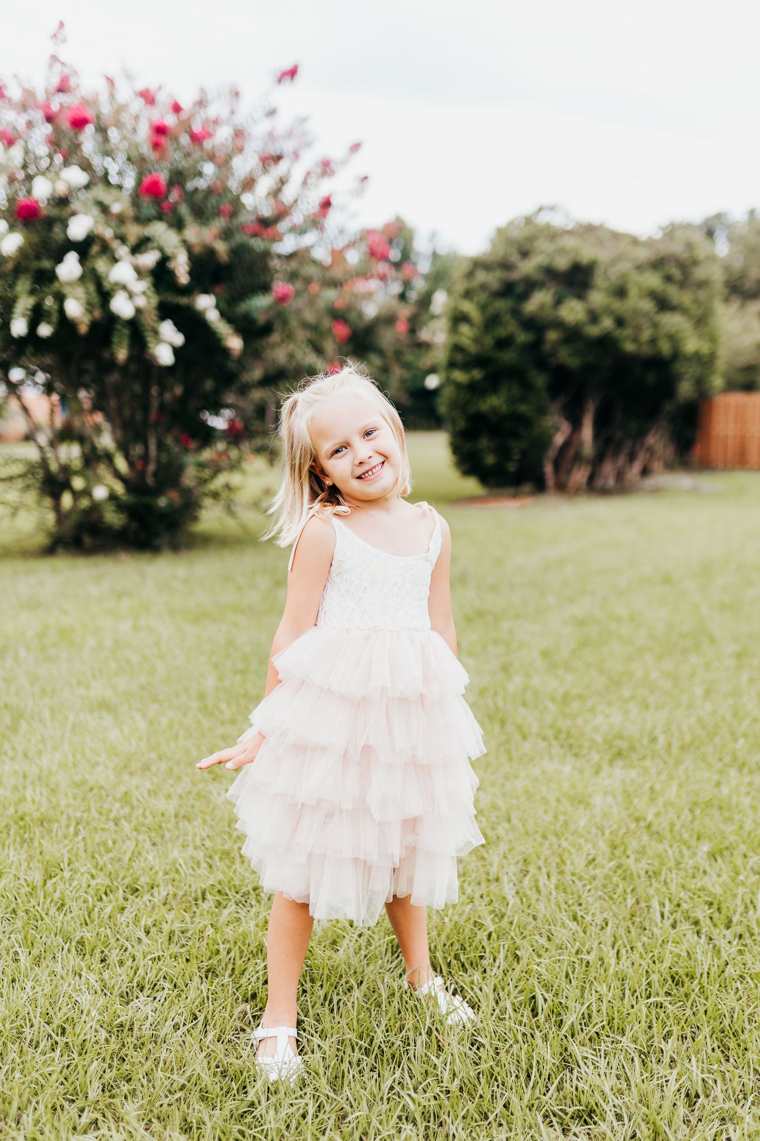 Young child in a white ruffled dress stands laughing joyfully on grass with flowering bushes in the background.