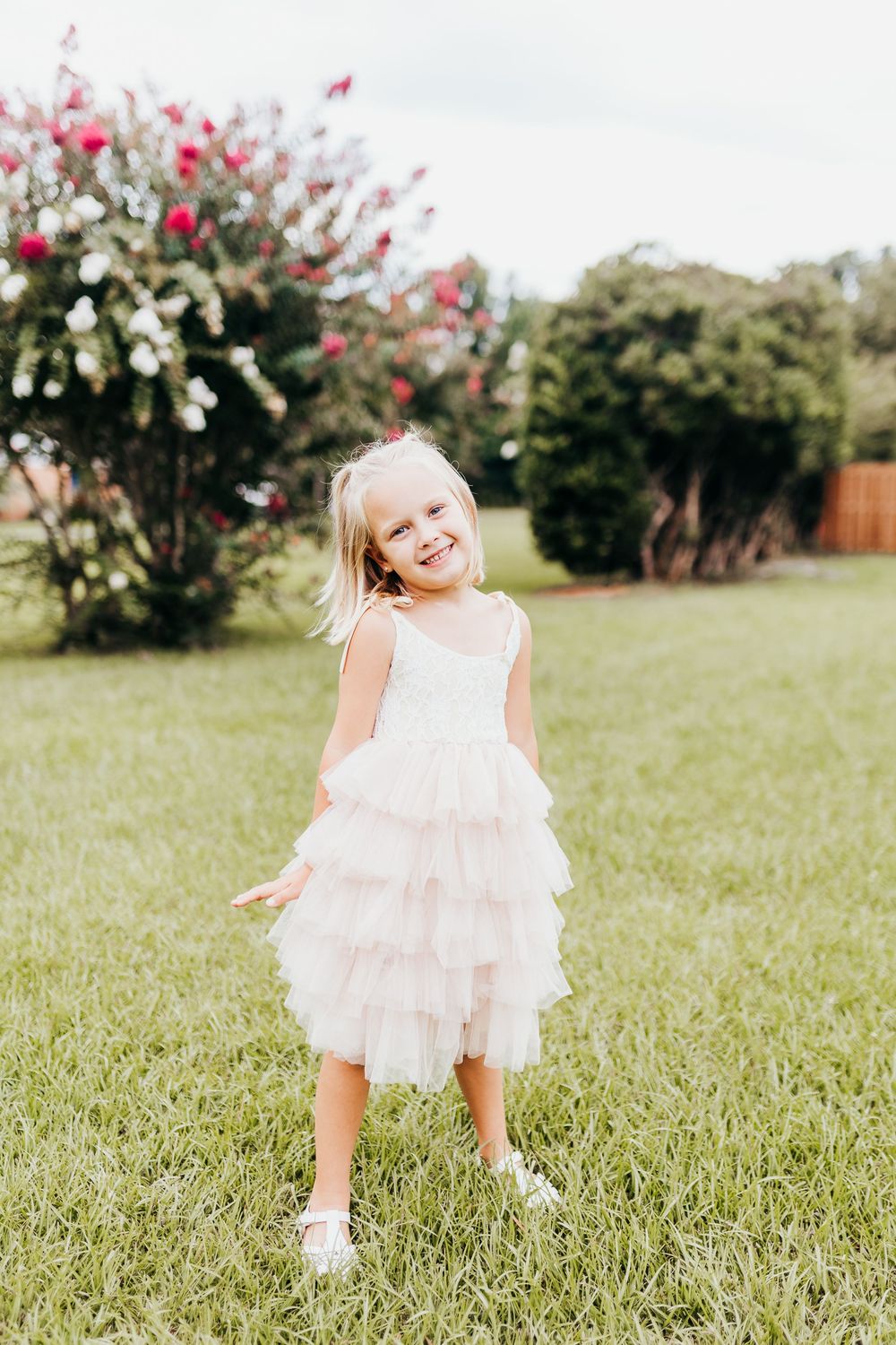Young child in a white ruffled dress stands laughing joyfully on grass with flowering bushes in the background.